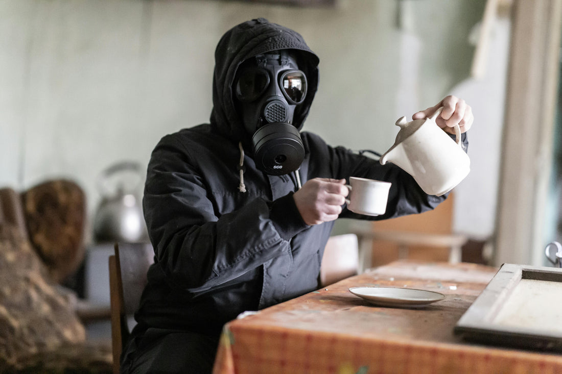 A person wearing a gas mask pouring coffee from a gooseneck kettle into a cup at a kitchen table, humorously illustrating the hidden toxins found in commodity coffee.