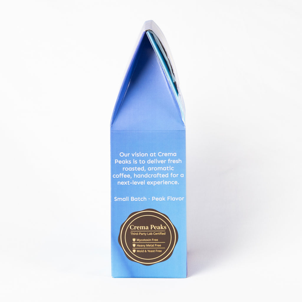 A tall, narrow blue coffee gift box with a pointed top, photographed on a clean white background. The front features white text and a circular gold and black logo for Crema Peaks.