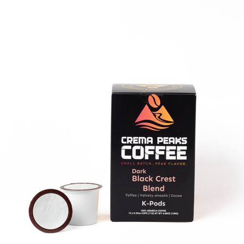 A box of Crema Peaks Coffee Dark Black Crest Blend K-Pods next to a single coffee pod capsule on a clean white background.