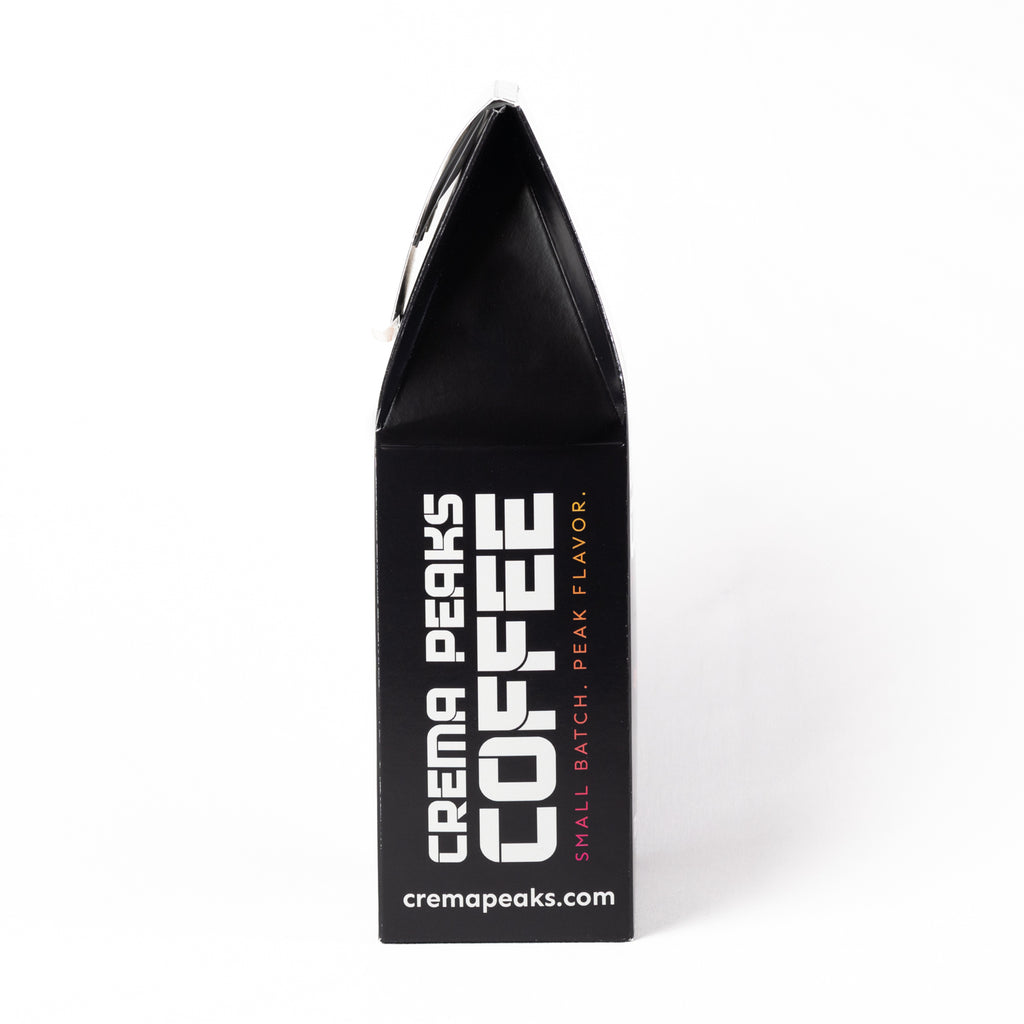 A black coffee bag package from Crema Peaks Coffee, standing upright on a white background. The packaging features bold white text reading 'CREMA PEAKS COFFEE' and smaller pink text reading 'SMALL BATCH, PEAK FLAVOR.