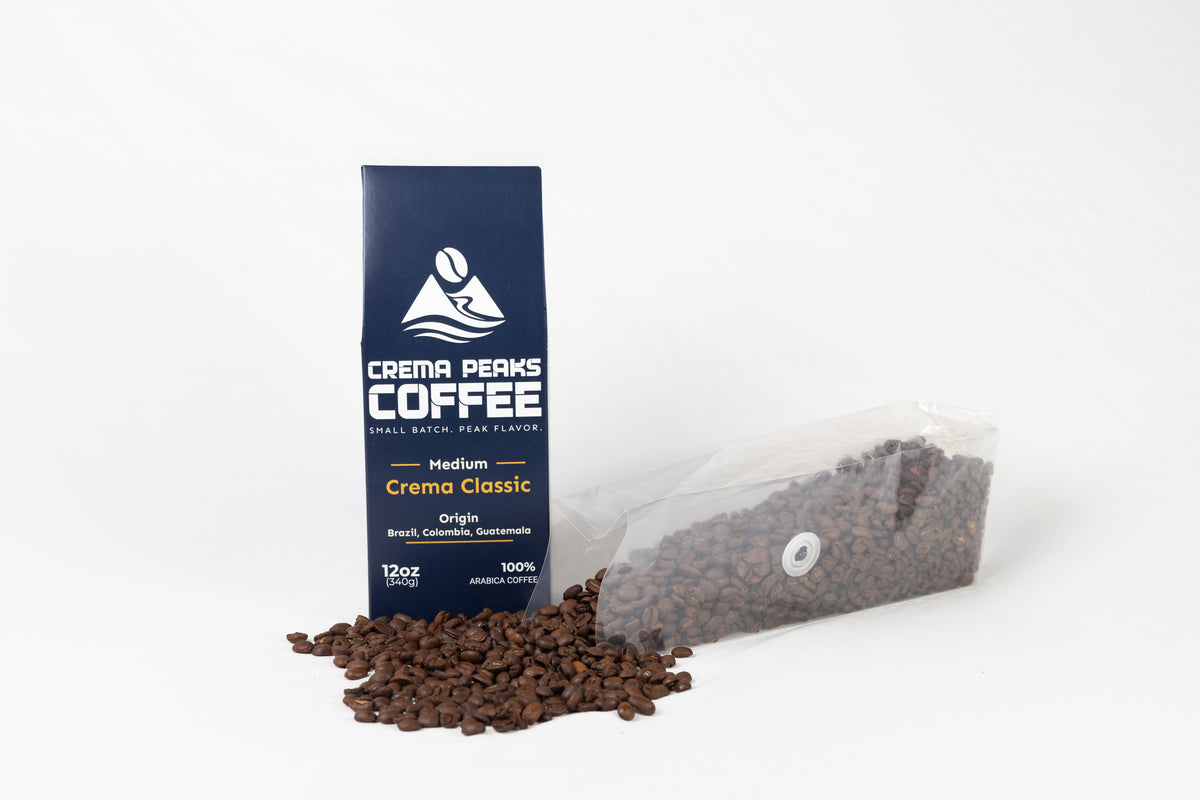 A bag of Crema Peaks Coffee Medium Crema Classic coffee stands upright next to a clear plastic bag filled with coffee beans, with a small pile of loose coffee beans in front, on a clean white background.