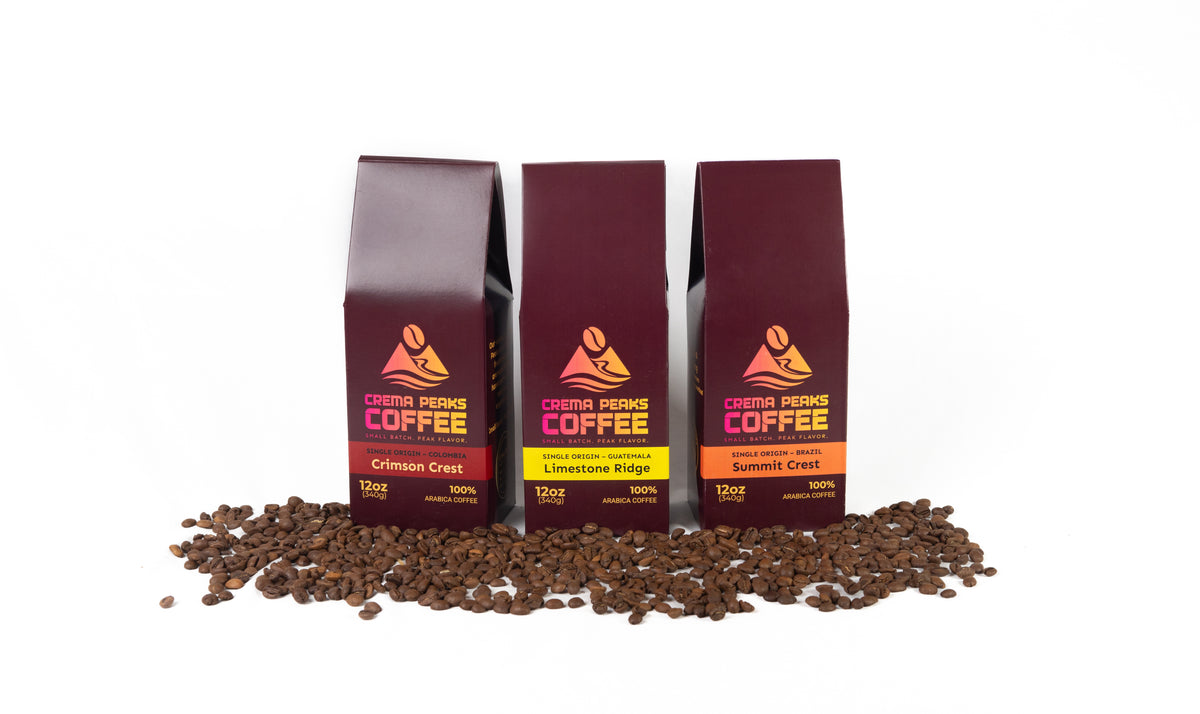 Three bags of Crema Peaks single origin Coffee arranged in a row on a white background, with coffee beans spilled in front. The bags are dark red-brown with yellow and orange labels, and the coffee beans are dark brown.