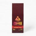 A tall, narrow box of Crema Peaks Coffee in the Crimson Crest single origin coffee, shown against a plain white background. The packaging is a deep maroon-brown color with a central graphic featuring a coffee bean, a mountain, and waves in yellow, orange, and pink, with text indicating it is a 12oz single-origin Colombian coffee.