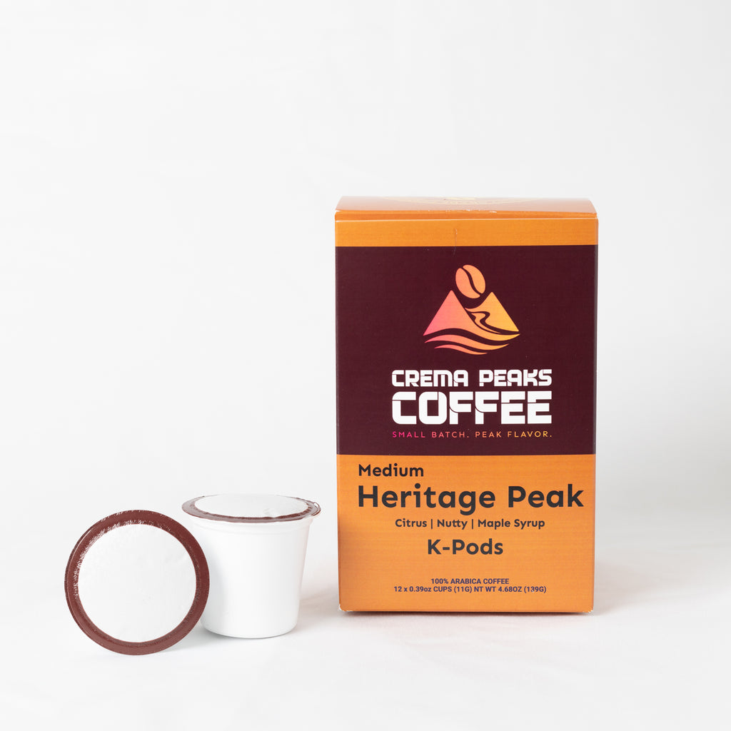 A box of Crema Peaks Coffee Medium Heritage Peak K-Pods coffee pods next to a single white coffee cup with a brown lid, on a clean white background.