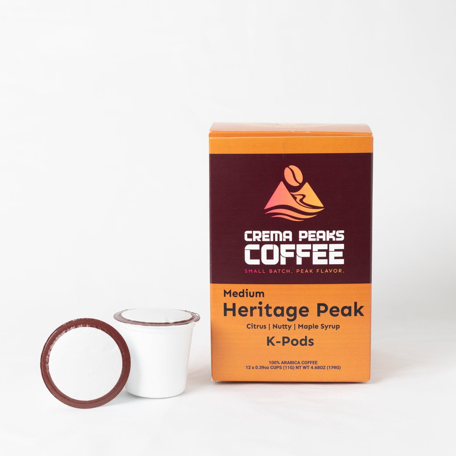 A box of Crema Peaks Coffee Medium Heritage Peak K-Pods coffee pods next to a single white coffee cup with a brown lid, on a clean white background.