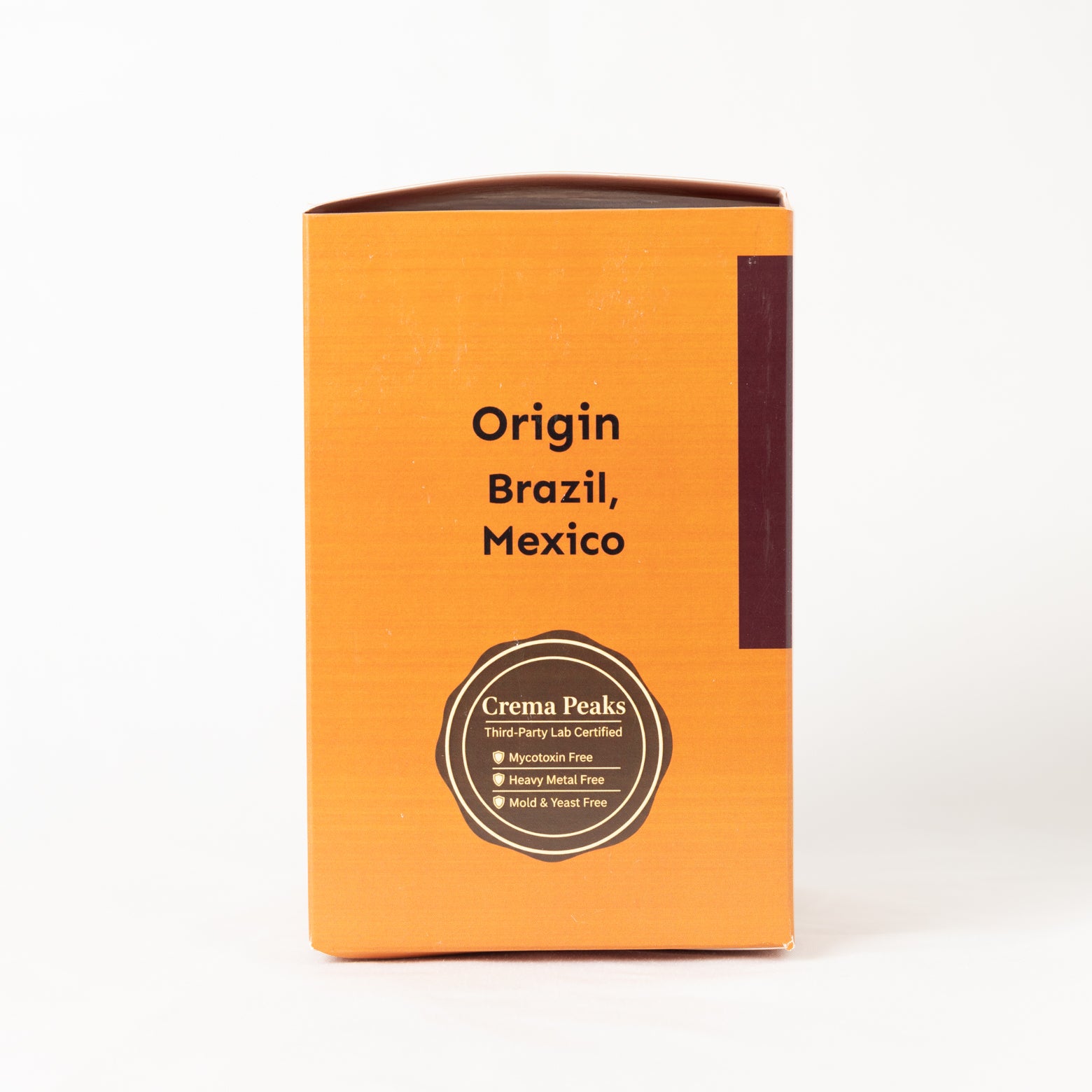 An orange rectangular product box with black text reading 'Origin Brazil, Mexico' and a circular 'Crema Peaks' certification seal near the bottom, photographed on a clean white background.
