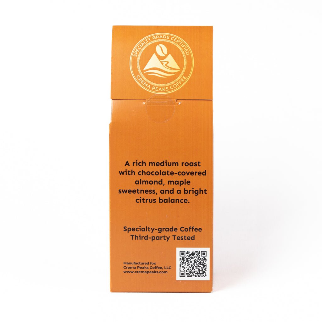 An orange cardboard box for Crema Peaks Coffee, showing the back side with product description text, a circular logo, and a QR code on a white background.