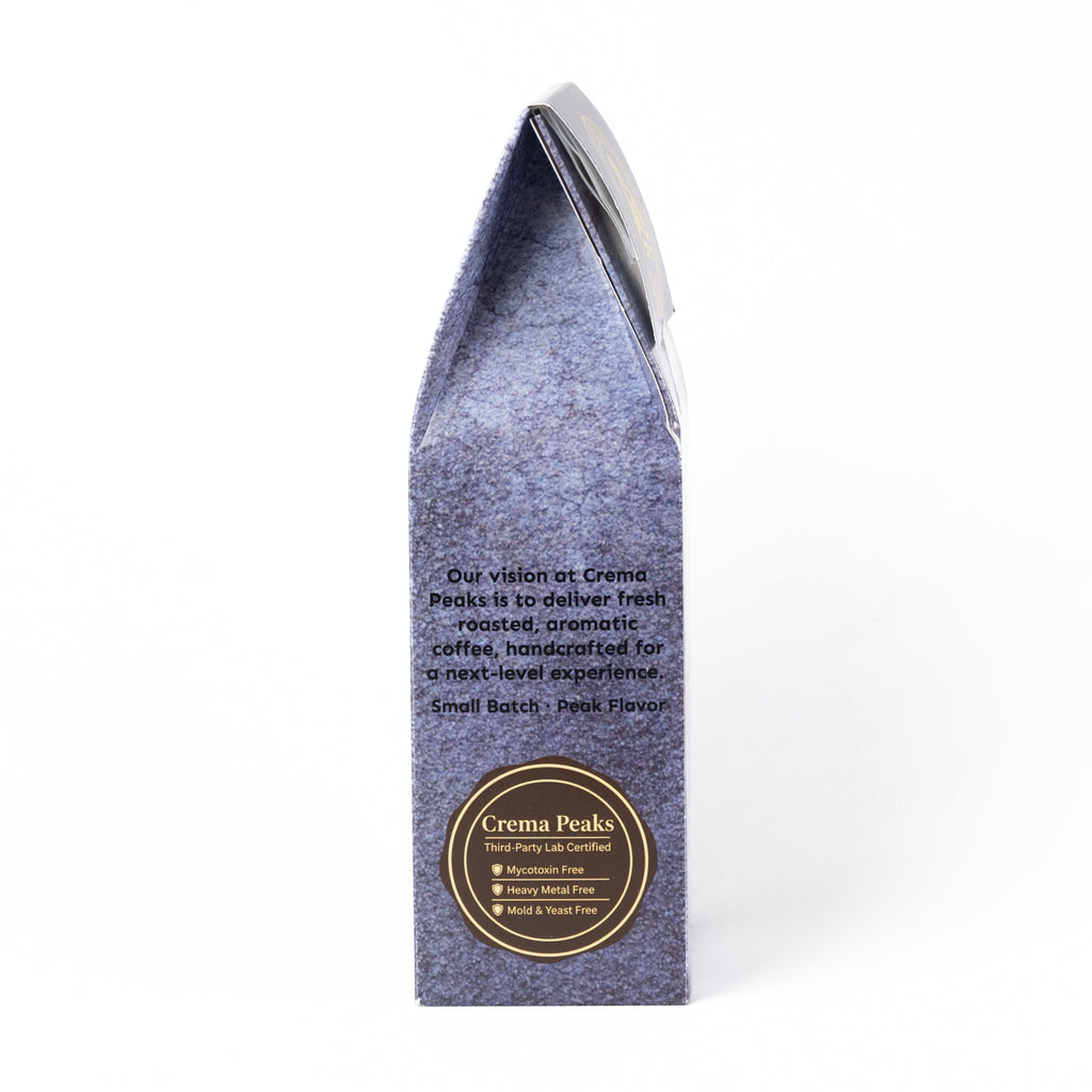 A single upright coffee bag in a textured periwinkle blue-gray paper sleeve with a black top flap, photographed on a clean white background. The front features a circular gold and black 'Crema Peaks' logo and descriptive text.