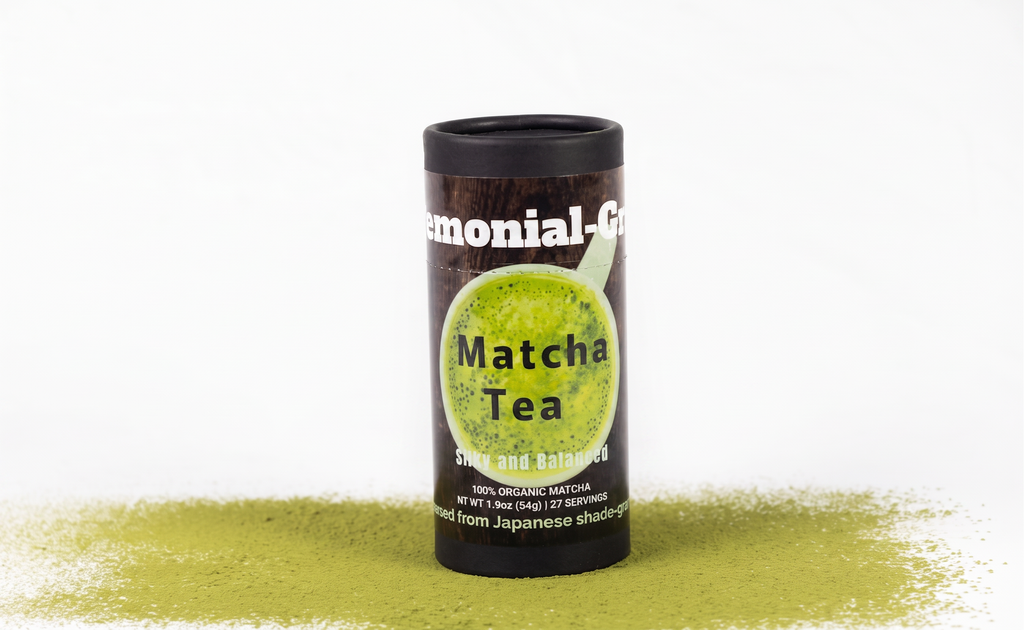 A black cylindrical container of organic ceremonial matcha tea with a green matcha tea illustration on the label, standing upright on a white background with matcha powder scattered at the base.