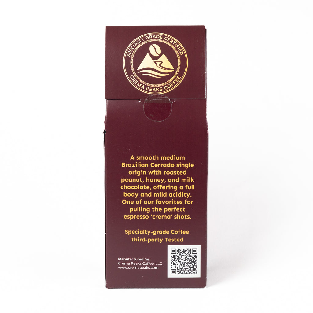 A tall, narrow dark burgundy coffee box shown upright on a white background, with gold text and a circular certified specialty-grade Coffee badge near the top. The front includes descriptive copy about a smooth medium Brazilian Cerrado single-origin coffee and a QR code near the bottom.