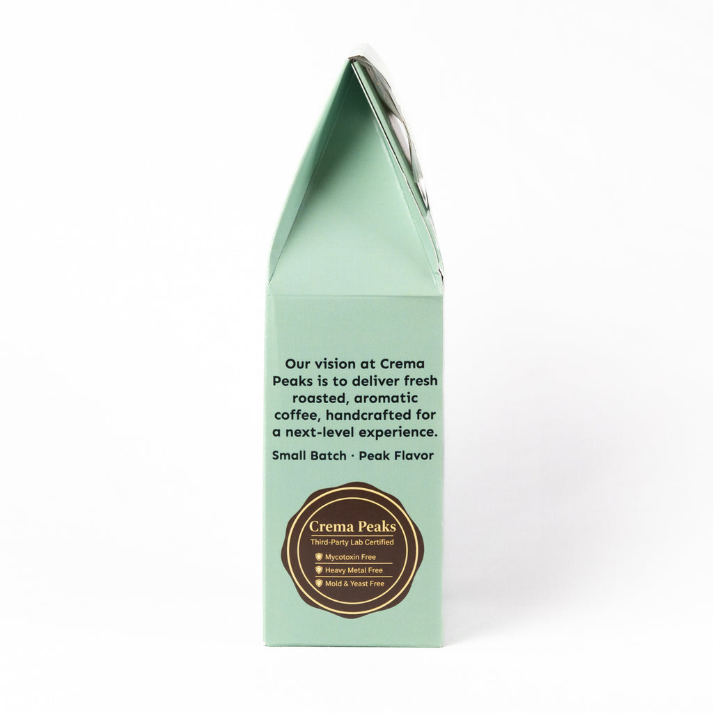 The back panel of a mint green paperboard coffee bag package by Crema Peaks, featuring black descriptive text and a brown circular logo seal on a white background.