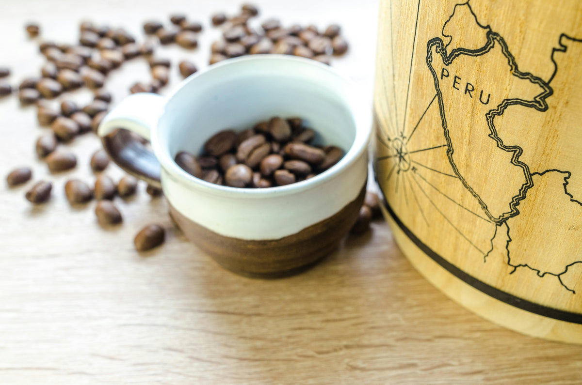 Cup filled with coffee beans next to a wooden container with a map design on a light wood surface.
